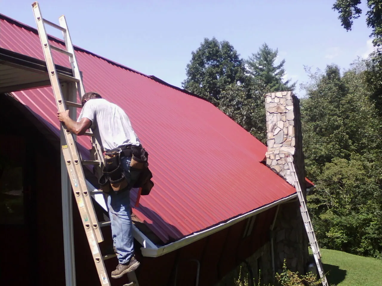 Roofer on a ladder beside a red metal roof and stone chimney — Soffit & Fascia Repair by Roofing Brothers in Little Sioux
